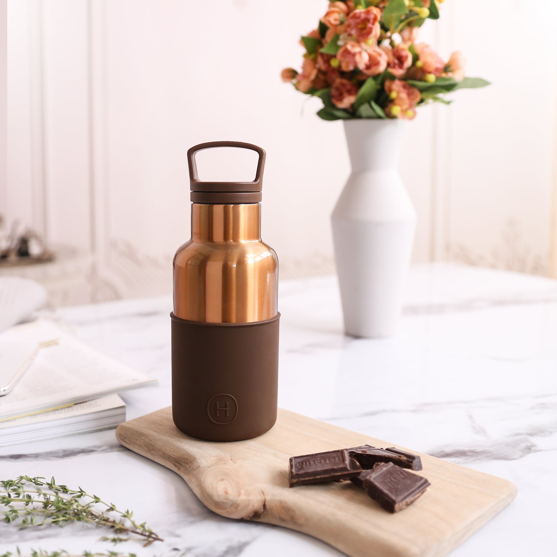 Bronze-colored insulated bottle on a wooden board with chocolate and flowers in the background