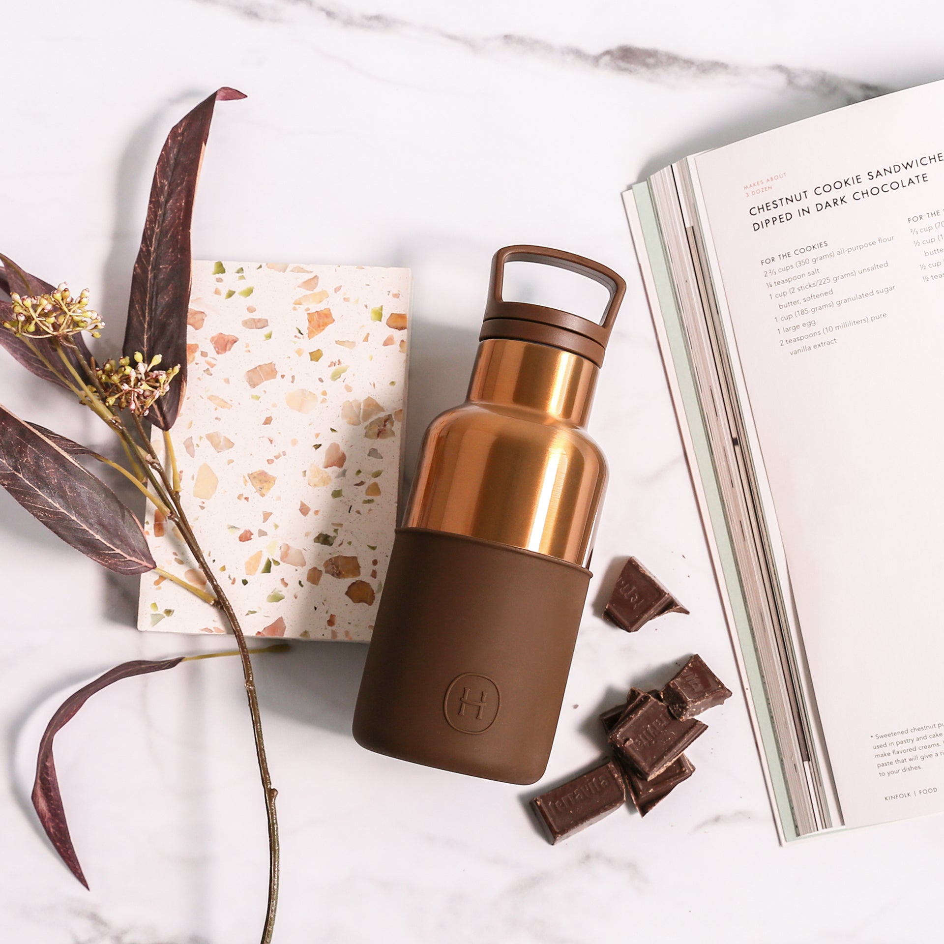 Brown and rose gold water bottle on a marble surface with chocolate and a book.
