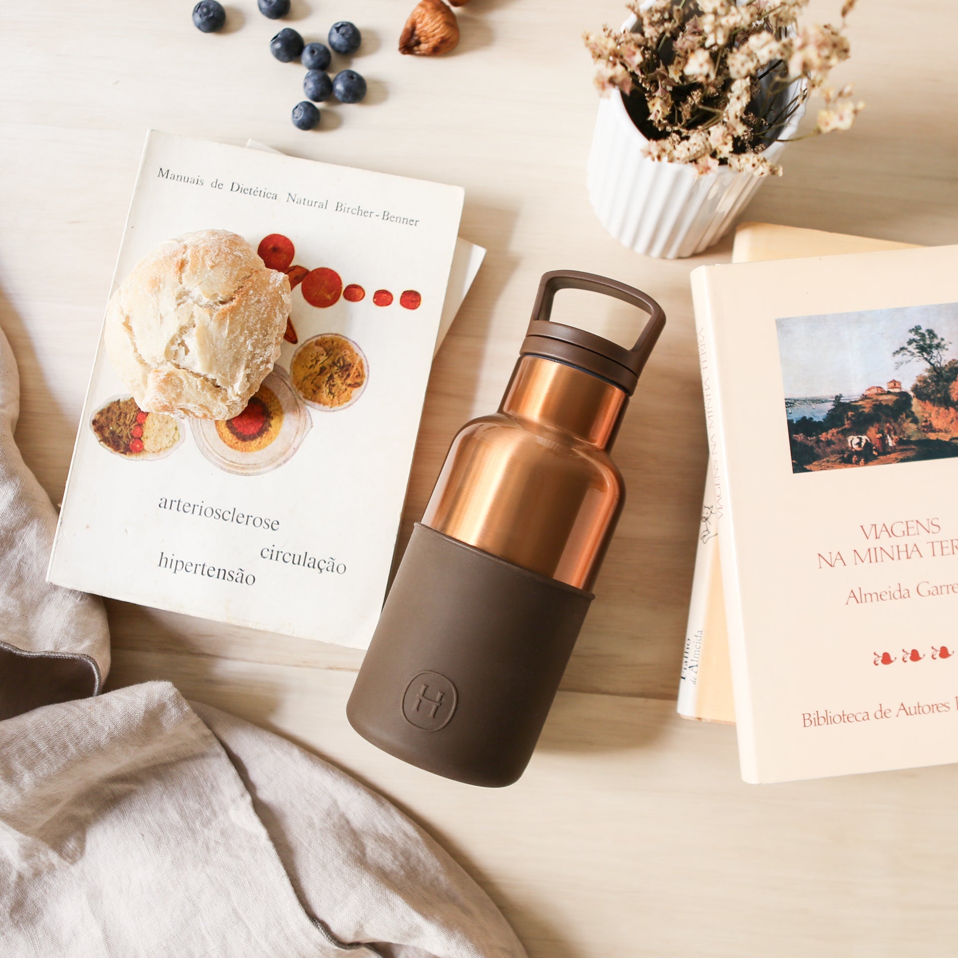 Bronze-colored water bottle with black base on a surface with books and blueberries.