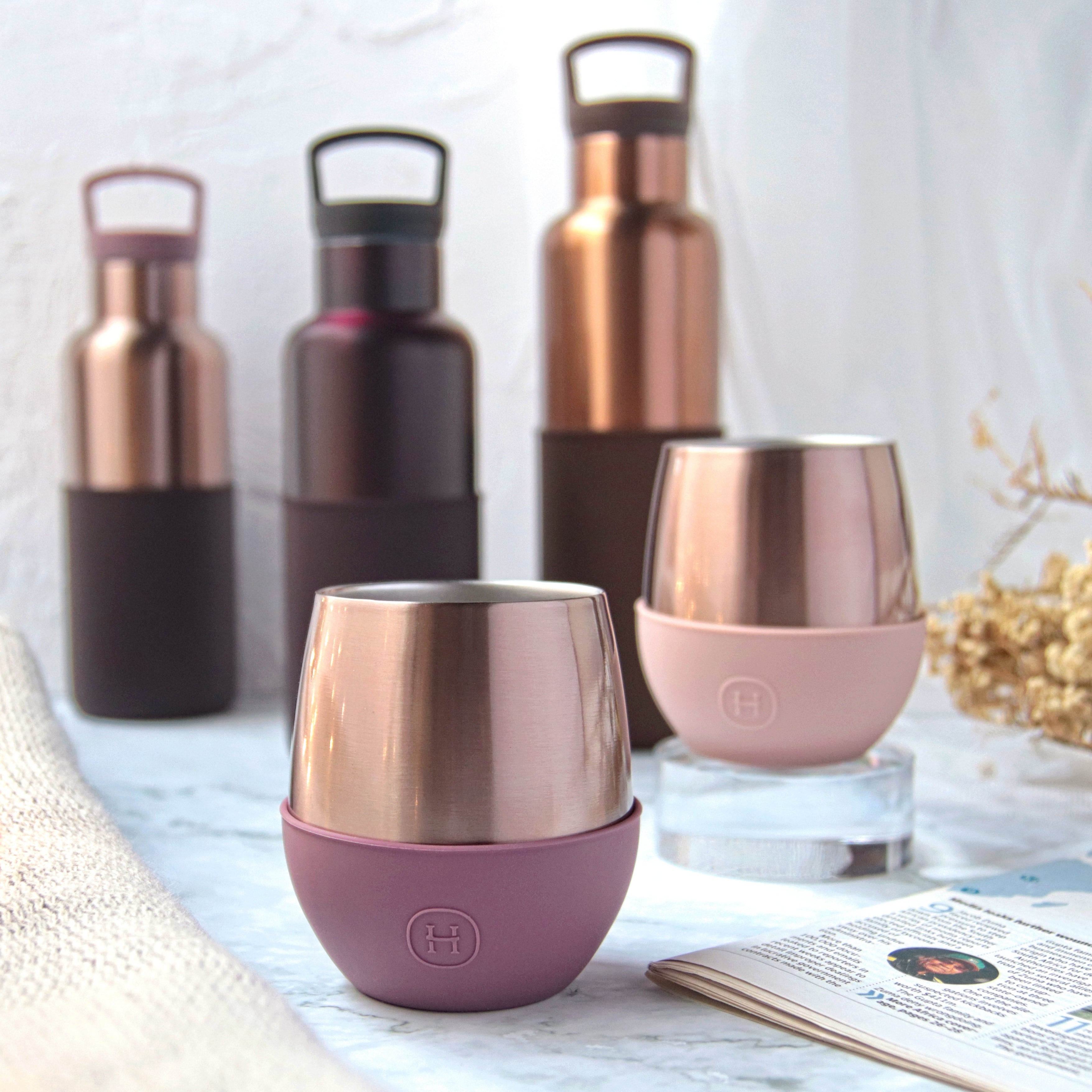 Set of rose gold and purple insulated cups on a table with a magazine and flowers.