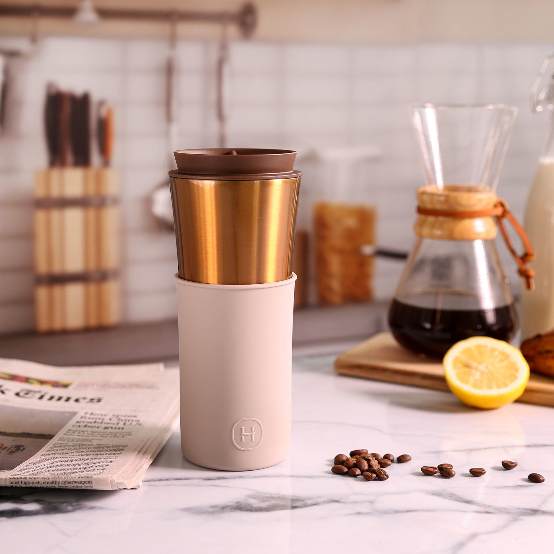 Travel mug with gold and beige design on a kitchen counter with coffee beans and a lemon.