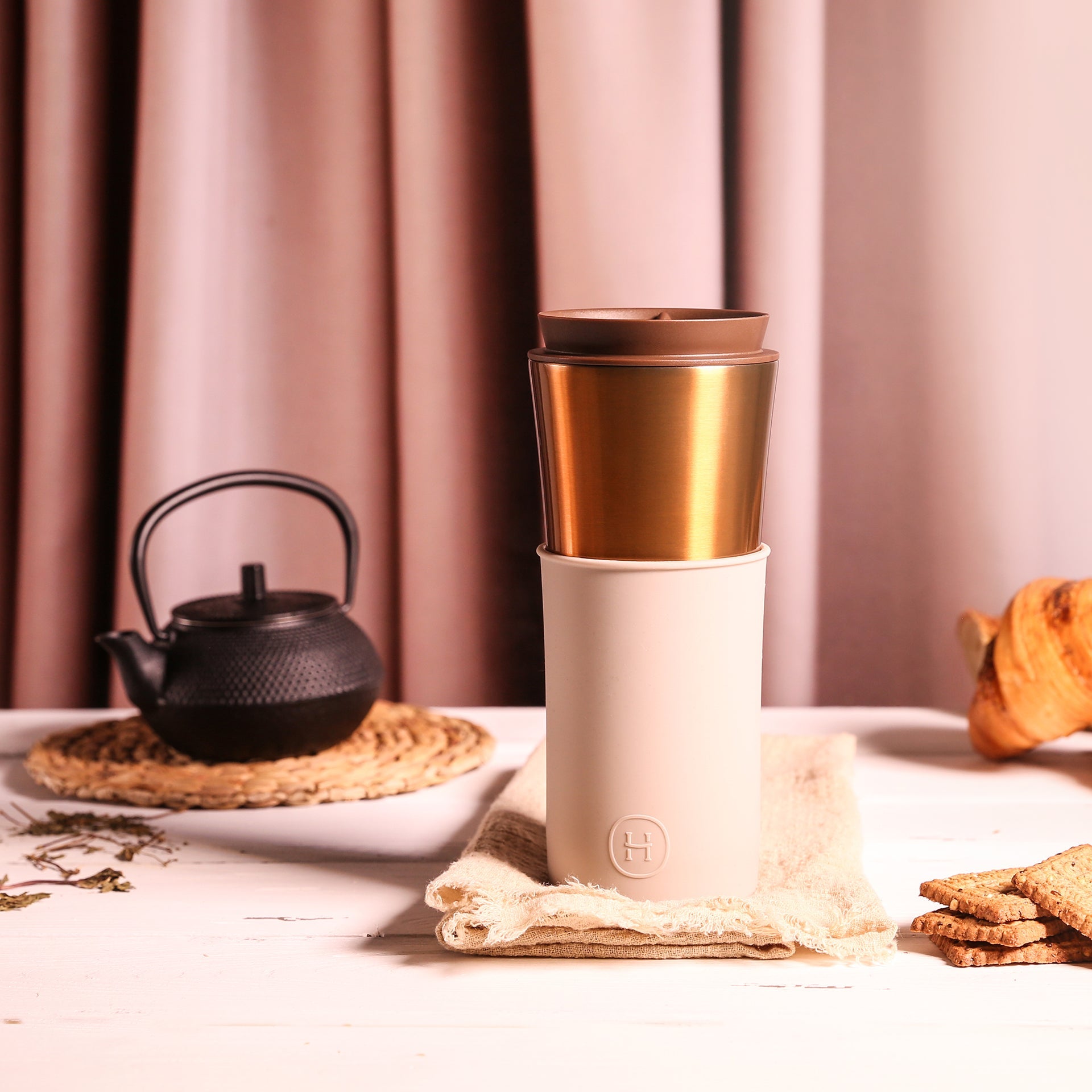 White and gold travel mug on a table with a teapot and cookies.