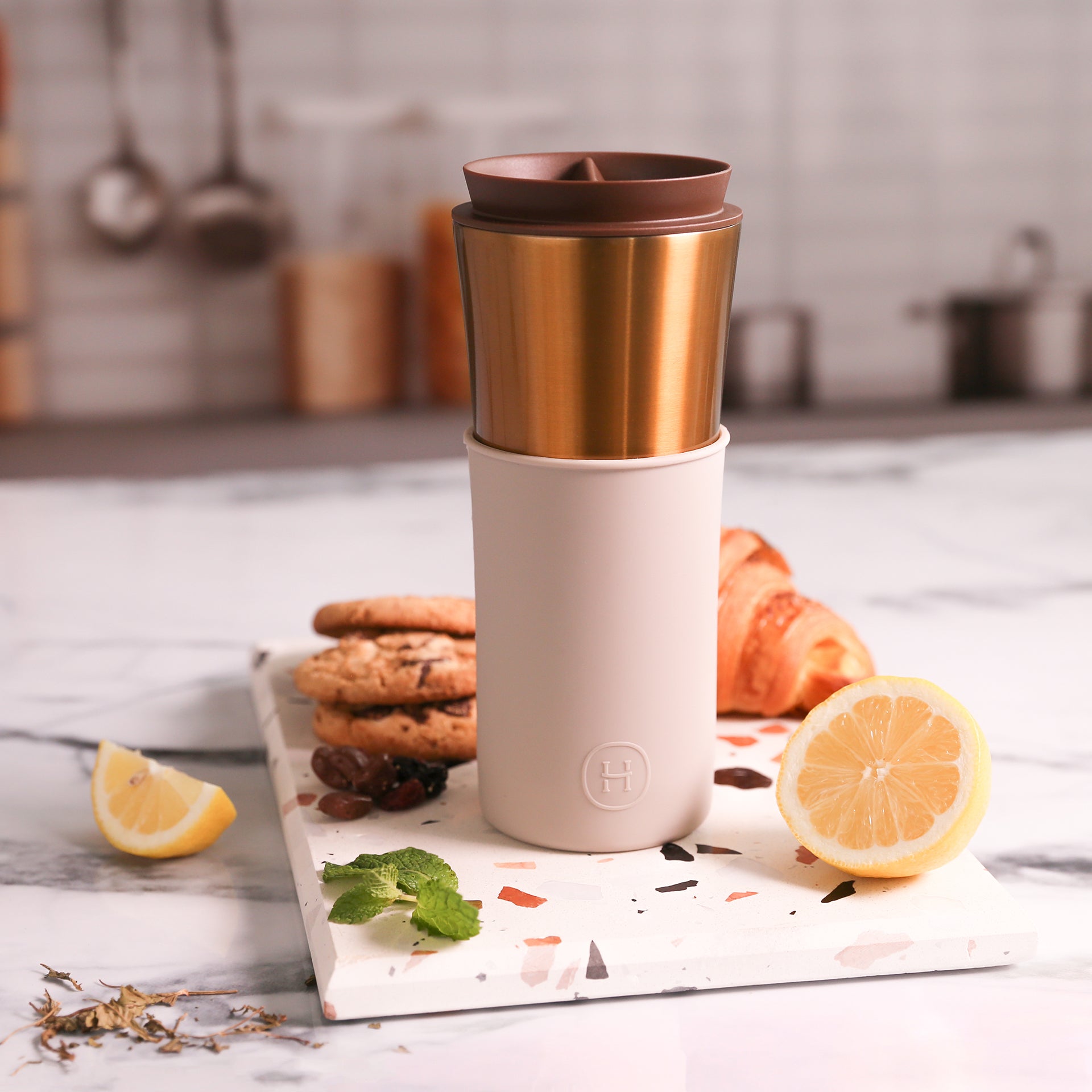 Travel mug with gold top and beige body on a kitchen counter with lemons, cookies, and a croissant.