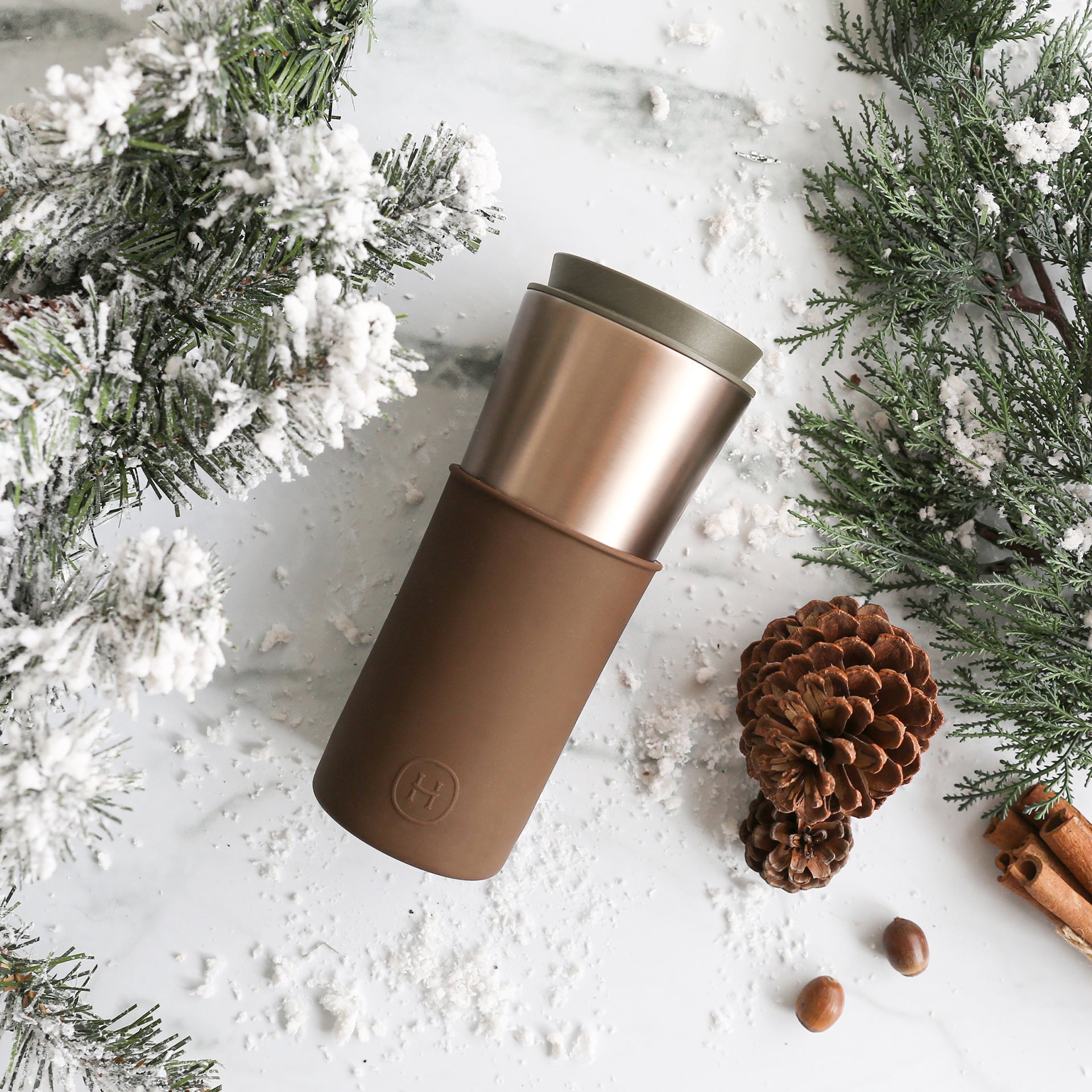 Brown travel mug with green lid on a snowy surface with pine cones and cinnamon sticks.