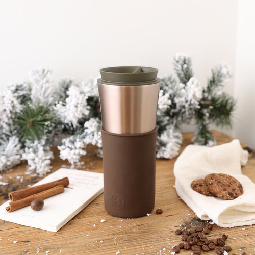 Brown and silver travel mug on a wooden surface with cookies and cinnamon sticks in the background.