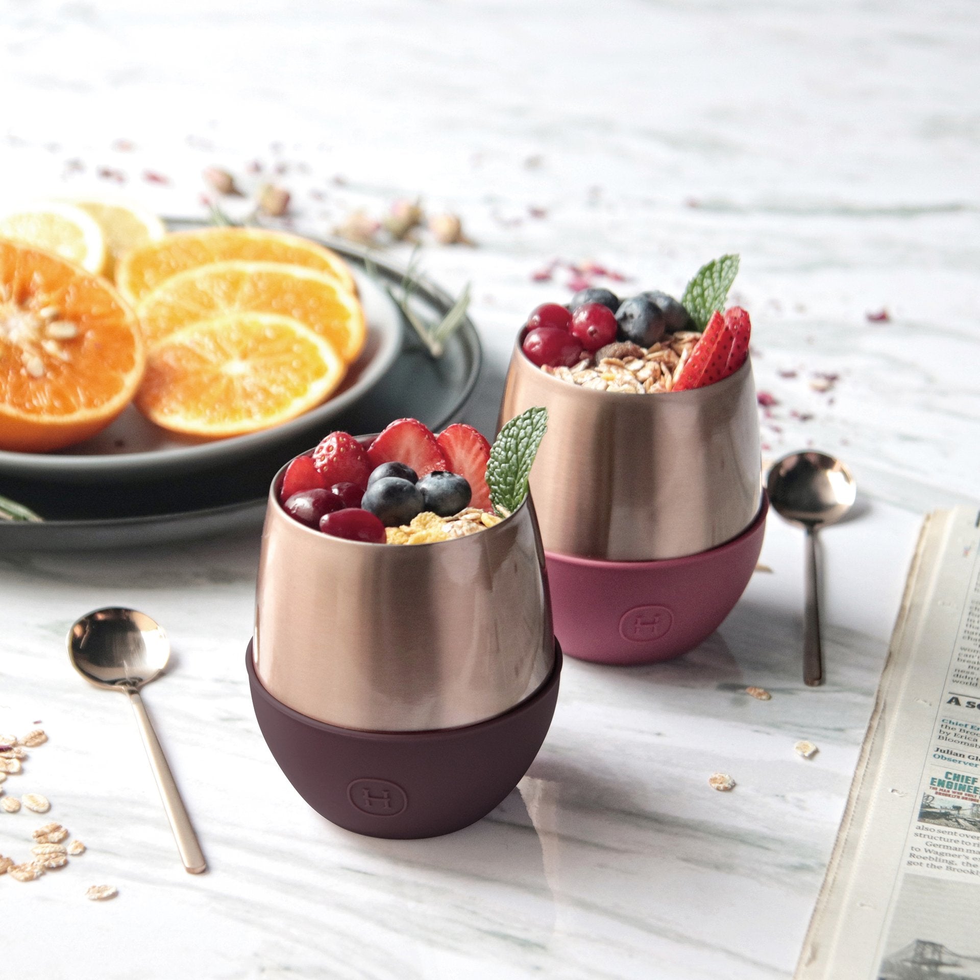 Two copper-colored tumblers filled with fruit and granola on a wooden table with oranges and a newspaper.