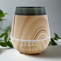 Wooden diffuser with black top on a light surface with green plants in the background